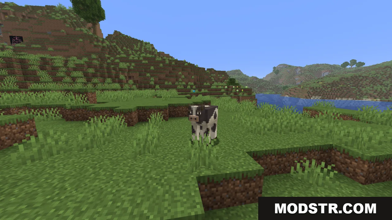 A spotted cow standing in a grassy field near a river and mountains